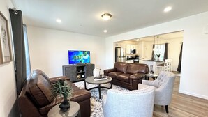 Ein modernes Wohnzimmer mit einem braunen Ledersofa, einem grauen Sessel und einem Couchtisch. Ein Flachbildfernseher ist an einer Kommode befestigt, und im Hintergrund ist eine Küche zu sehen.