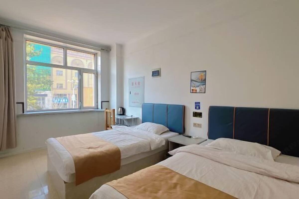A hotel room with two beds, a window with curtains, a small table with a kettle, and a wall-mounted TV.