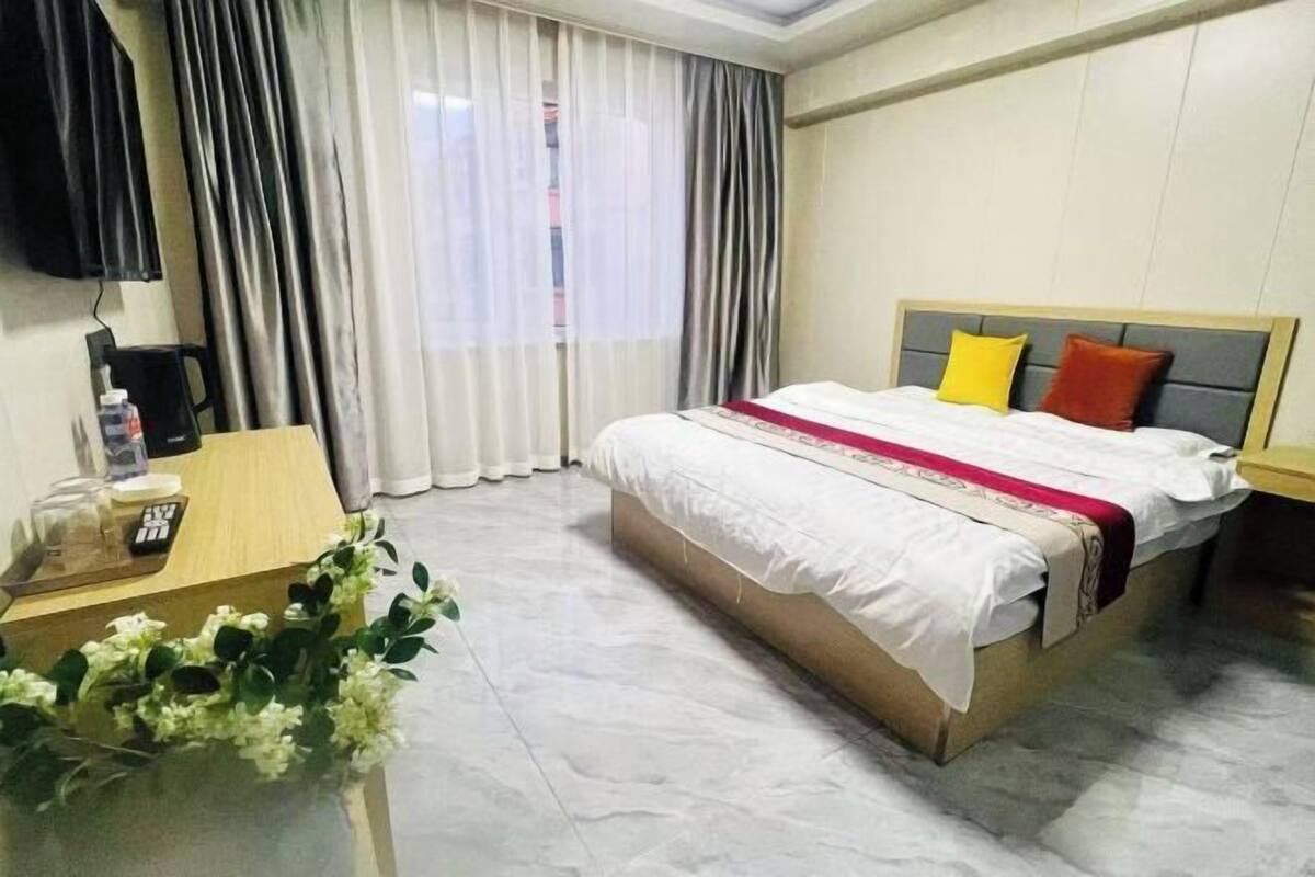 A hotel room with a bed, a desk with a telephone, a small table with a vase of flowers, and a window with curtains.