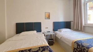 Une chambre d’hôtel avec deux lits, une petite table, une poubelle, une fenêtre avec des rideaux et un seul tableau accroché au mur.