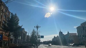 Une large rue bordée de bâtiments des deux côtés, un ciel dégagé et un soleil éclatant.