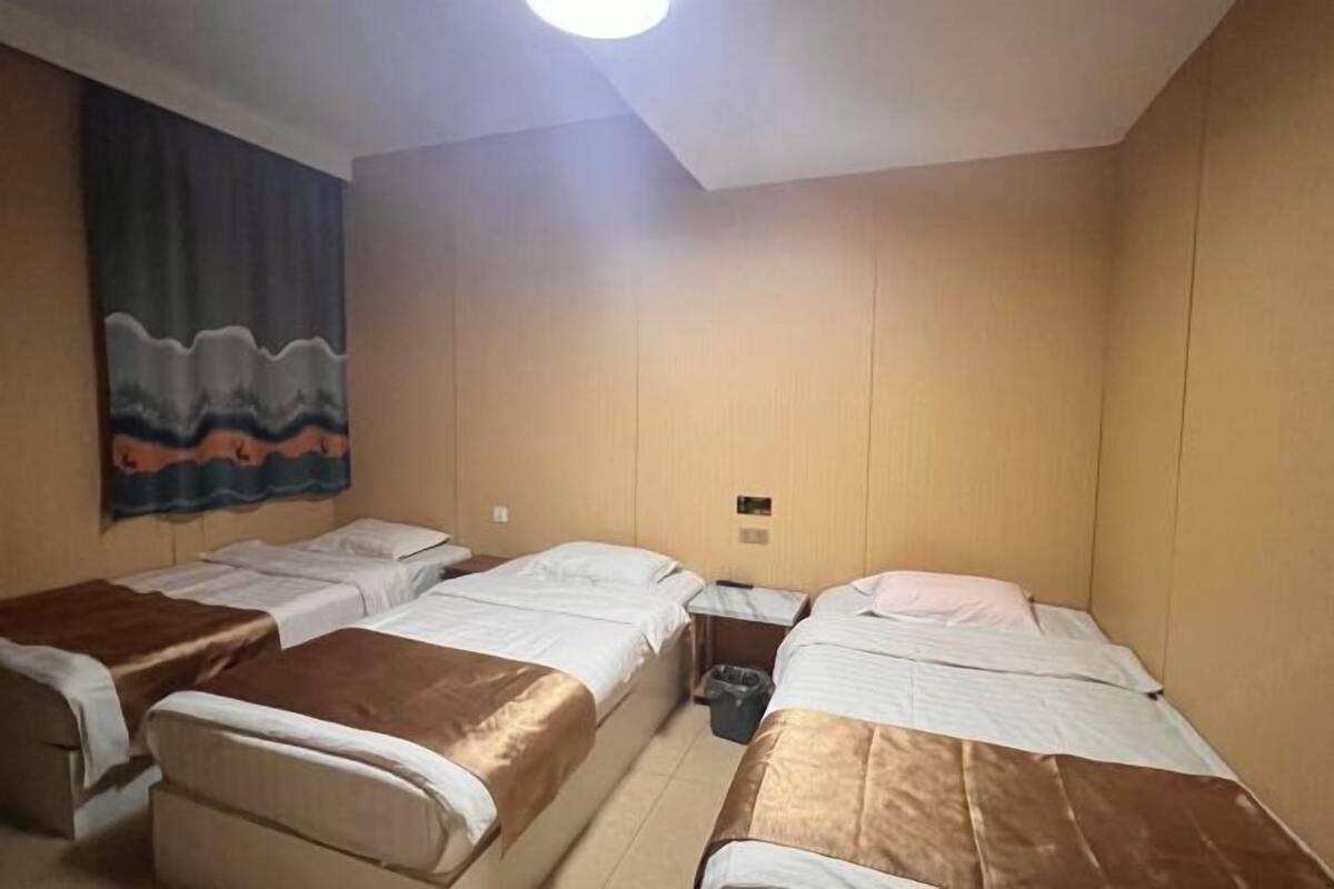 A room with four single beds arranged in a row, a ceiling light, and a wall with a colorful tapestry.