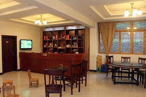 Un vestíbulo de hotel con una recepción, un televisor, una zona de comedor con una mesa redonda y sillas, y un ventanal con cortinas.