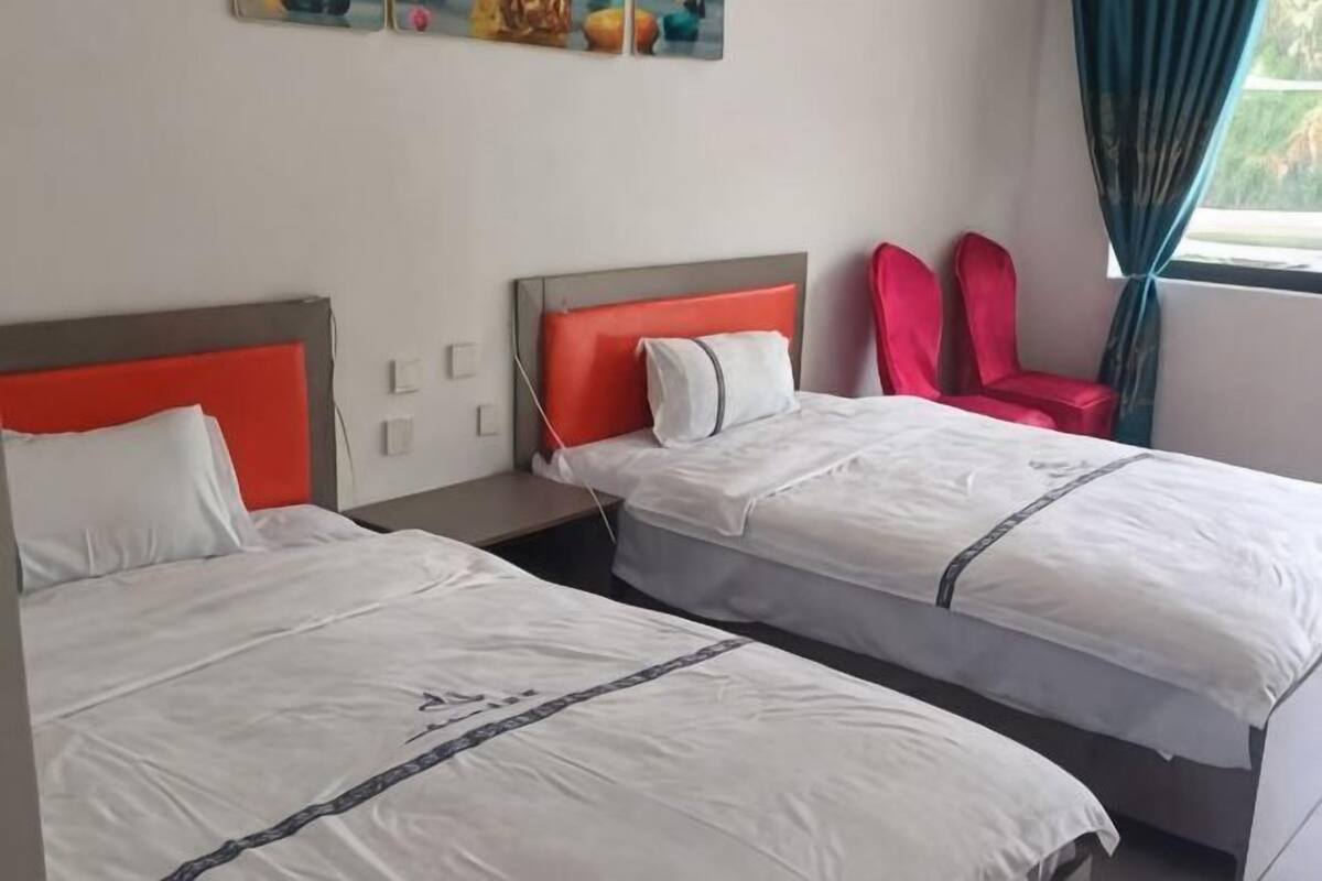 Une chambre d’hôtel avec deux lits, un fauteuil rouge et une fenêtre avec des rideaux.