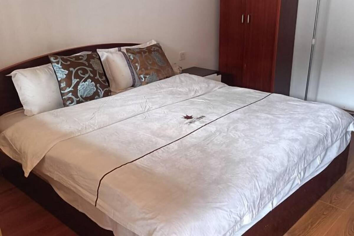 Un lit bien fait, avec un couvre-lit blanc et des coussins décoratifs, une tête de lit en bois et une armoire en bois dotée d’un miroir.