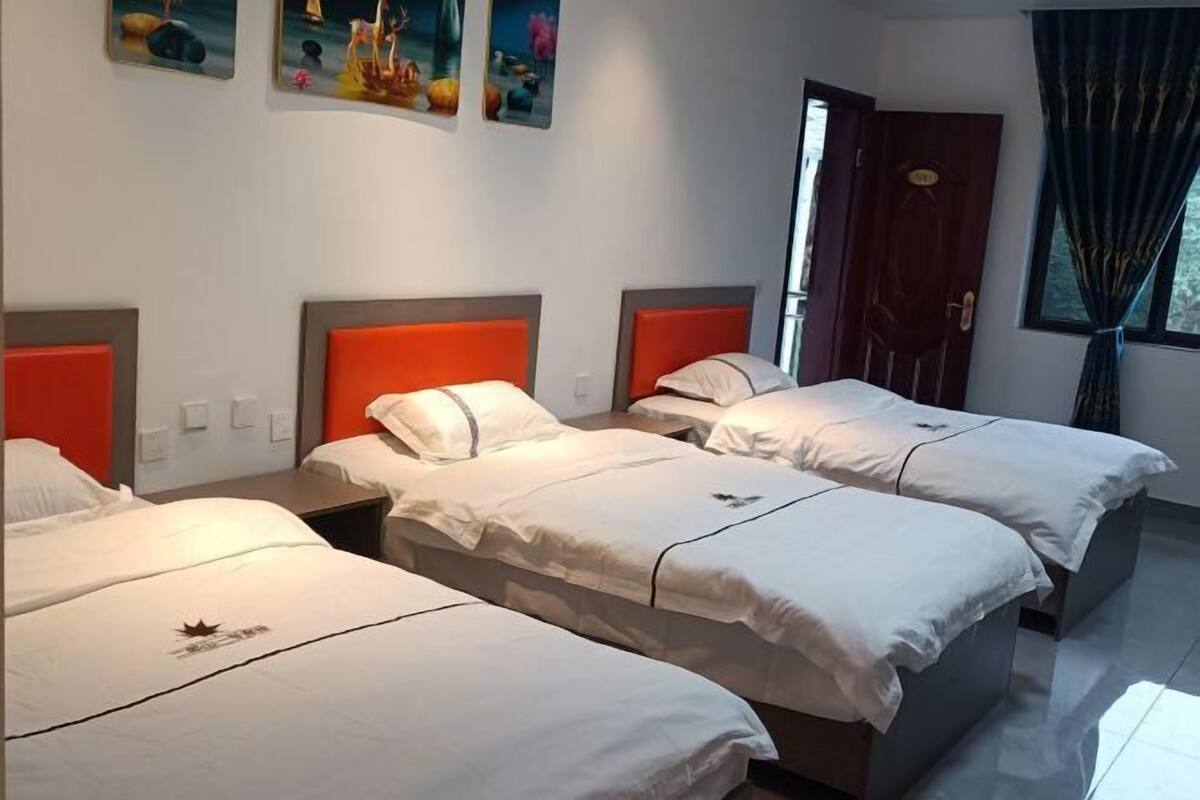 Une chambre d’hôtel avec trois lits, chacun recouvert d’une courtepointe blanche et doté d’une tête de lit orange. Des tableaux sont accrochés au mur au-dessus des lits, et une porte avec un rideau se trouve à droite.