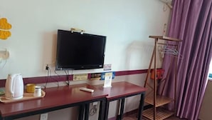 A hotel room with a TV mounted on the wall, a wooden desk with a tray holding a kettle, a remote control, and a small container. There is a wooden shelf unit with a hanging rack and a purple curtain in the background. - Kangchao Hotel, Yueqing (Yueqing)