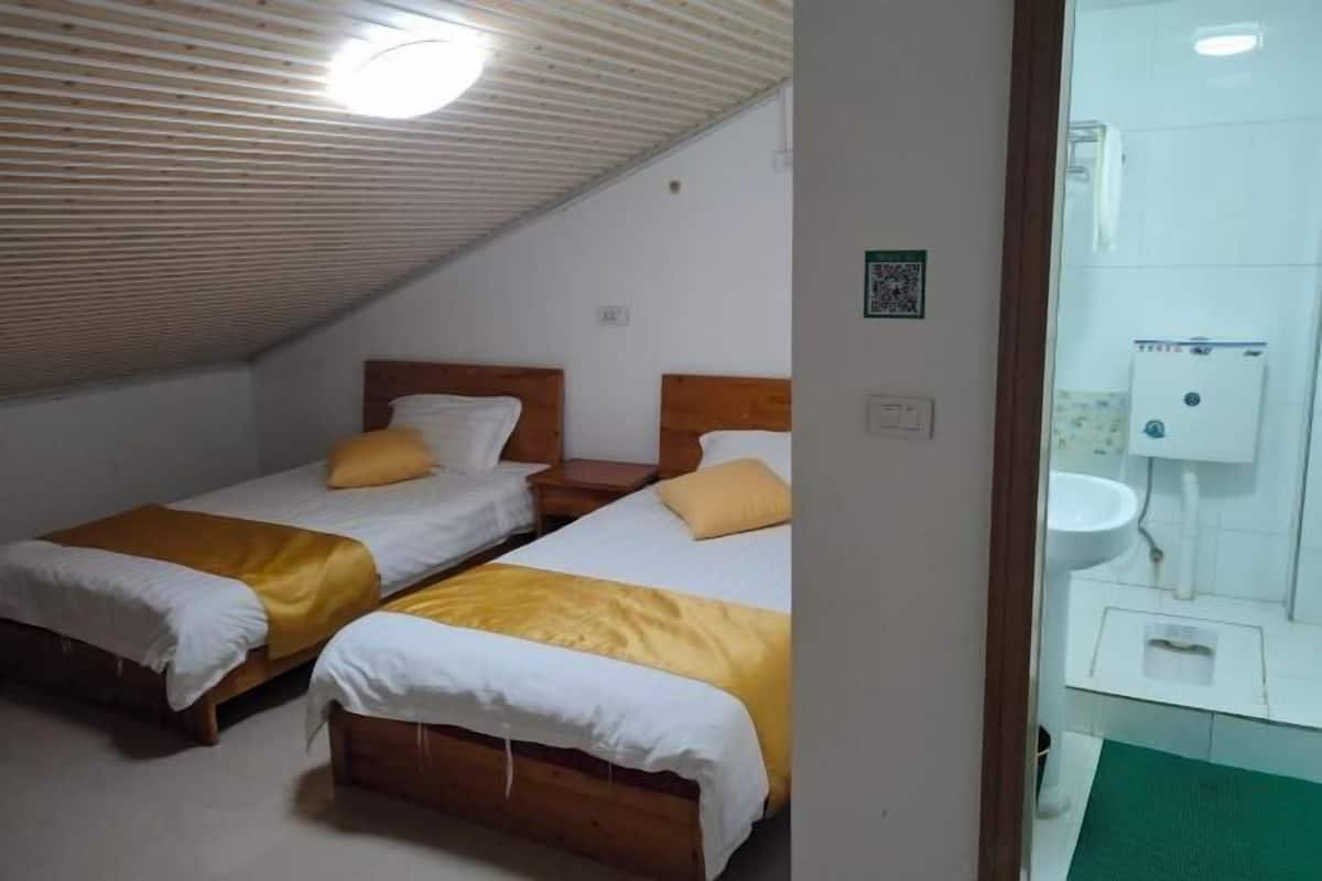 Une chambre avec deux lits en bois, une petite table de chevet, un seul luminaire et une vue sur une salle de bain équipée d’un lavabo et de toilettes.