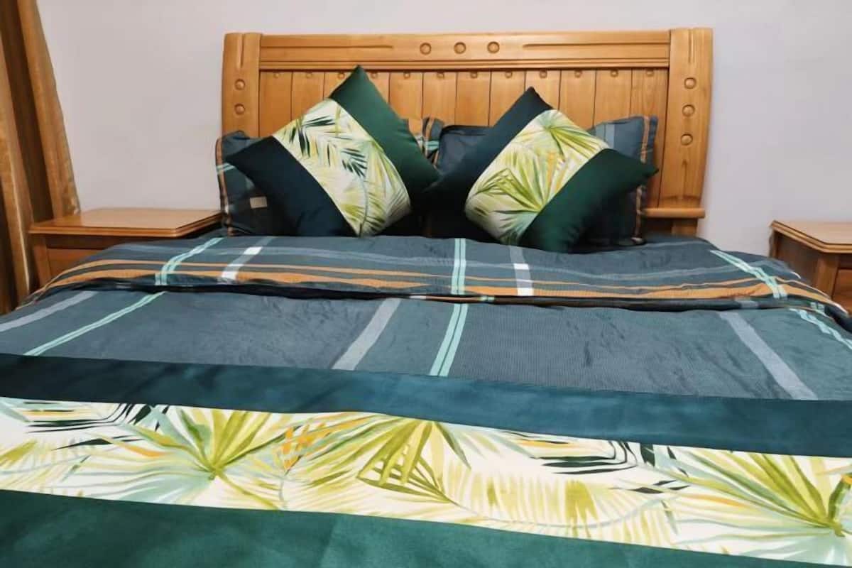 Un lit avec une tête de lit en bois, deux oreillers verts et un oreiller vert foncé, et une couvre-lit ornée d’un motif de feuilles tropicales.