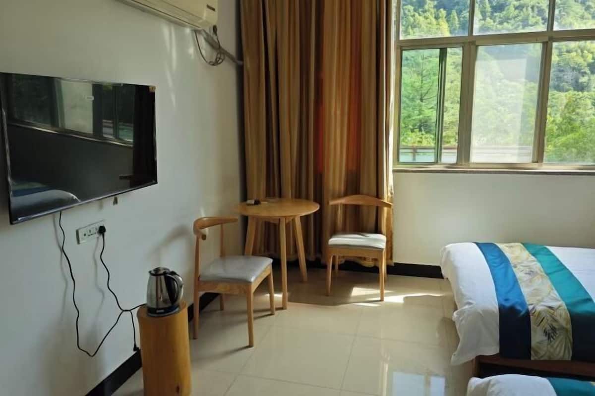 Une chambre d’hôtel avec un lit, un téléviseur fixé au mur, une petite table avec des chaises et une fenêtre donnant sur un paysage verdoyant.