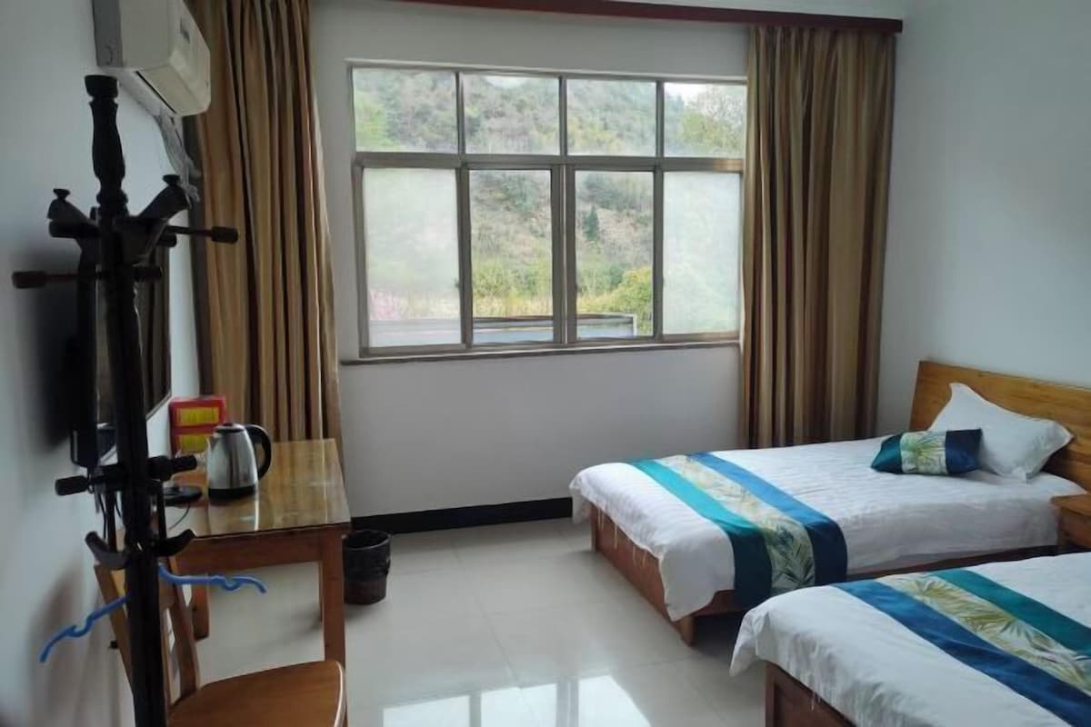 Habitación de hotel con dos camas, una ventana con cortinas, una mesa de madera con hervidor y una silla.