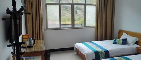 A hotel room with two beds, a window with curtains, a wooden table with a kettle, and a chair.