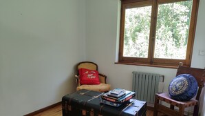 Una habitación con piso de madera, una silla con cojín rojo, un cojín azul con bordados dorados y una mesa con una caja de Scrabble y una caja de rompecabezas.