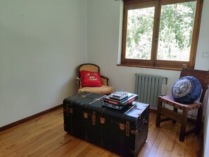 Une pièce avec un plancher en bois, une chaise avec un coussin rouge, un coussin bleu orné de broderies dorées, et une table sur laquelle se trouvent une boîte de Scrabble et une boîte de jeux de puzzle.
