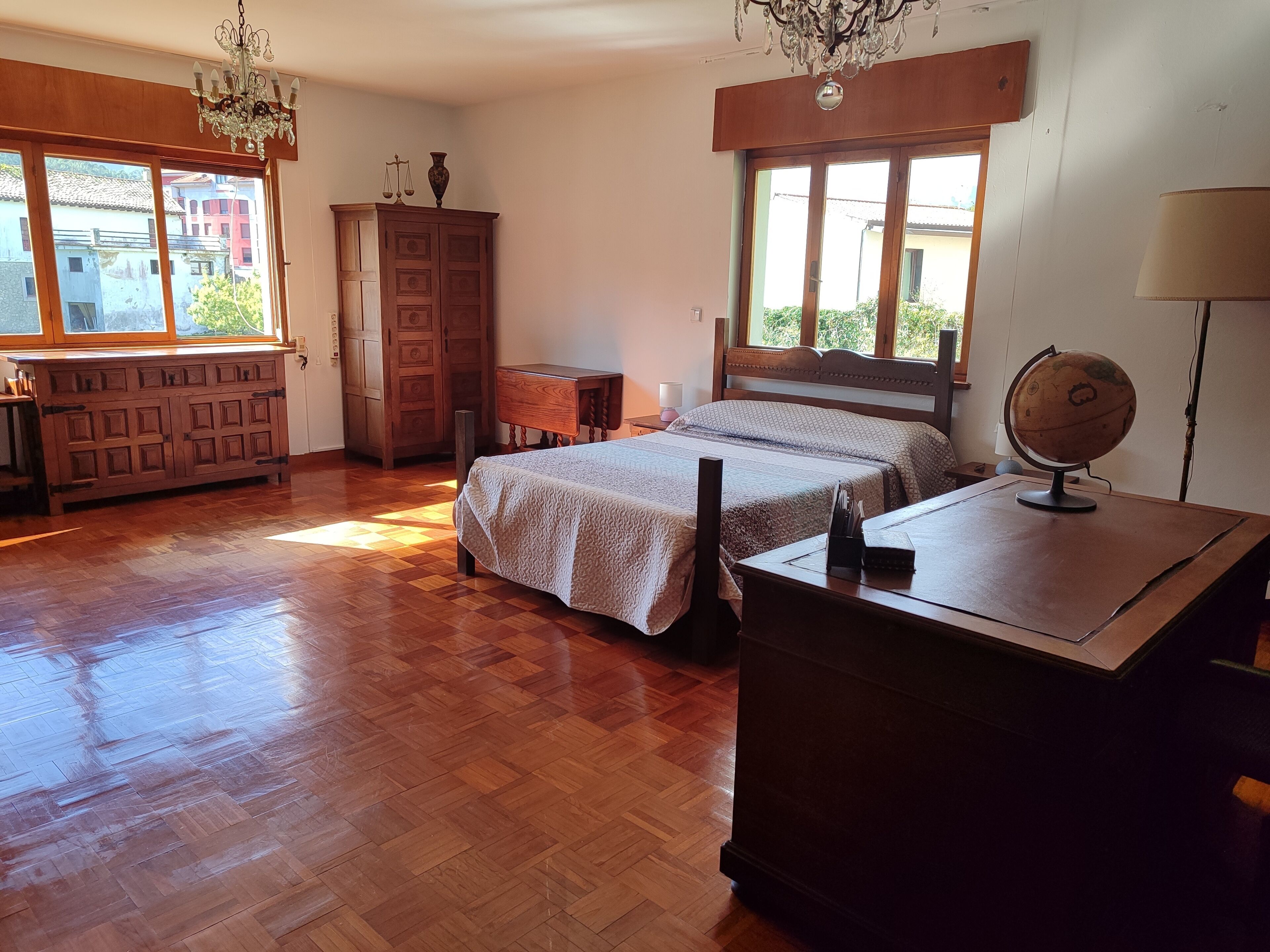 A bedroom with a wooden floor, a bed with a patterned bedspread, a wooden dresser, a desk with a globe, and a window with a view of buildings.