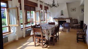 Comedor con una mesa preparada para cuatro personas, rodeada de sillas de madera. Hay una chimenea con repisa de piedra y una araña cuelga del techo.