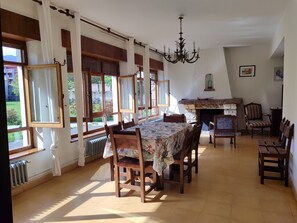 Uma sala de jantar com uma mesa posta para quatro pessoas, cercada por cadeiras de madeira. Há uma lareira com manto de pedra, e um lustre pendurado no teto.
