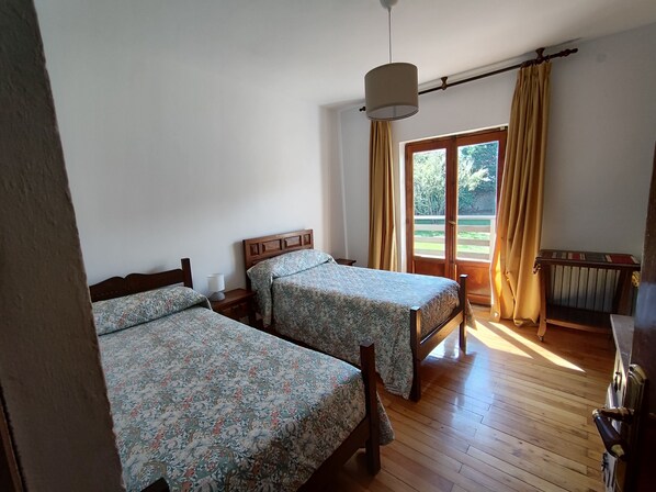 Une chambre avec deux lits en bois, recouverts d’une literie à motifs floraux, un plancher en bois, une fenêtre avec des rideaux et un radiateur.