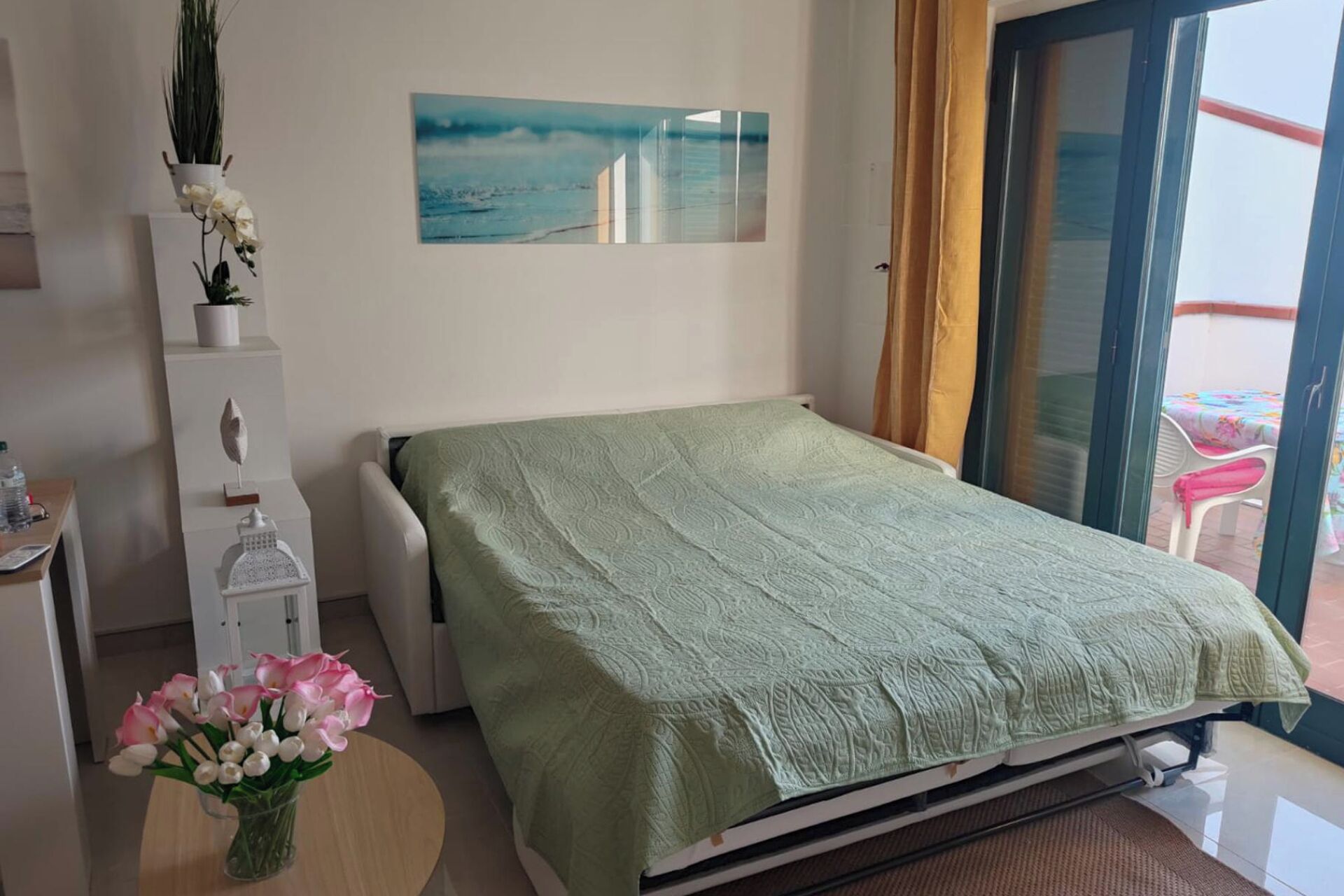 Une chambre à coucher avec un lit, une petite table sur laquelle est posé un vase de fleurs, une plante en pot et un tableau accroché au mur.