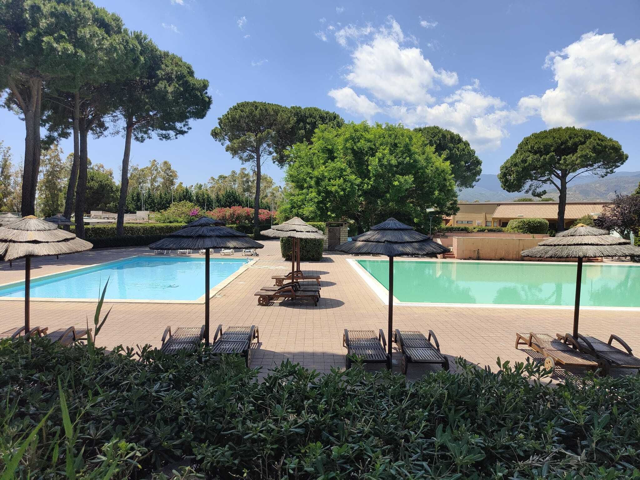 Un espace piscine comprenant deux piscines, entouré de chaises longues et de parasols, avec des arbres et un bâtiment en arrière-plan.