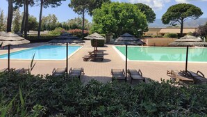 Area piscina con due vasche, circondata da lettini e ombrelloni, con alberi e un edificio sullo sfondo.