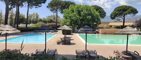 A pool area with two pools, surrounded by lounge chairs and umbrellas, with trees and a building in the background.