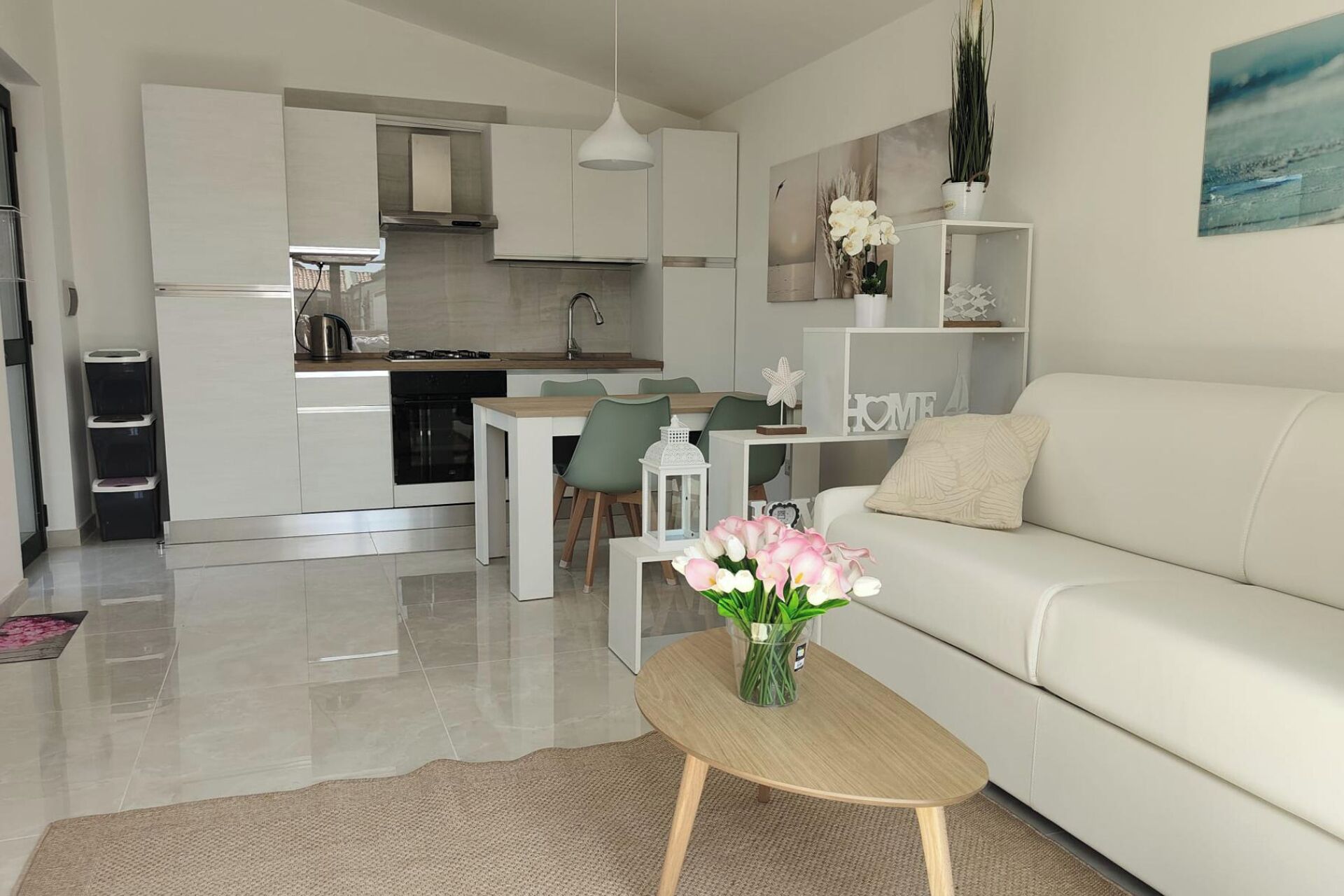 Moderni olohuone, jossa on valkoinen sohva, kukilla koristeltu sohvapöytä, ruokailutila tuoleineen sekä taustalla näkyvä keittiö.