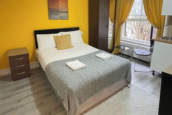 A hotel room with a bed, a small desk, a chair, a window with curtains, and a painting on the wall.