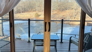 A wooden deck with a table and chairs, a glass door with curtains, and a view of a natural landscape. - Beautiful Modern mountain getaway overlooking water with fountain. (Joplin)