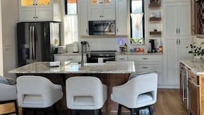 A modern kitchen with white cabinetry, a central island with bar stools, a stainless steel refrigerator, and a microwave. - Beautiful Modern mountain getaway overlooking water with fountain. (Joplin)