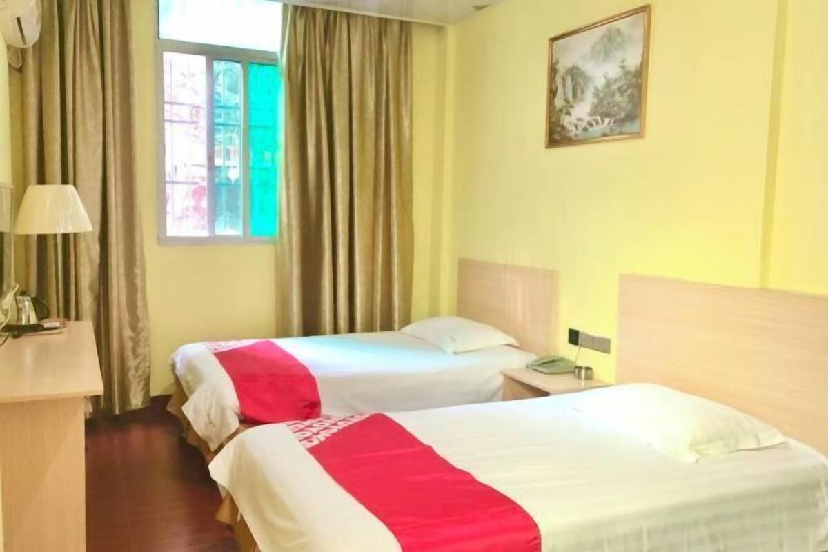 A hotel room with two beds, a desk, a lamp, a painting on the wall, and a window with curtains.