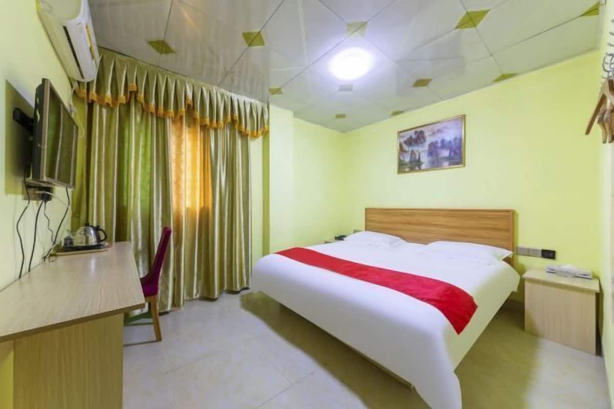 A hotel room with a bed, a desk, a chair, curtains, a painting, and a ceiling light.