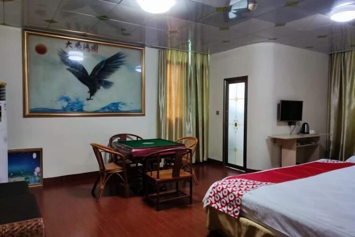 A hotel room with a bed, a desk with a chair, a table with chairs, a painting on the wall, a television, and a window with curtains.