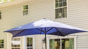 Een terras met een tafel en stoelen onder een grote parasol, een barbecue en een schuifdeur naar een achtertuin.