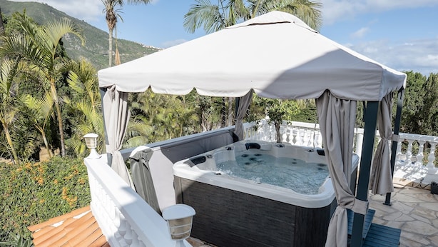 Een hottub onder een wit dak, omringd door een witte hek en palmbomen, met een stenen terras en een houten vlonder.