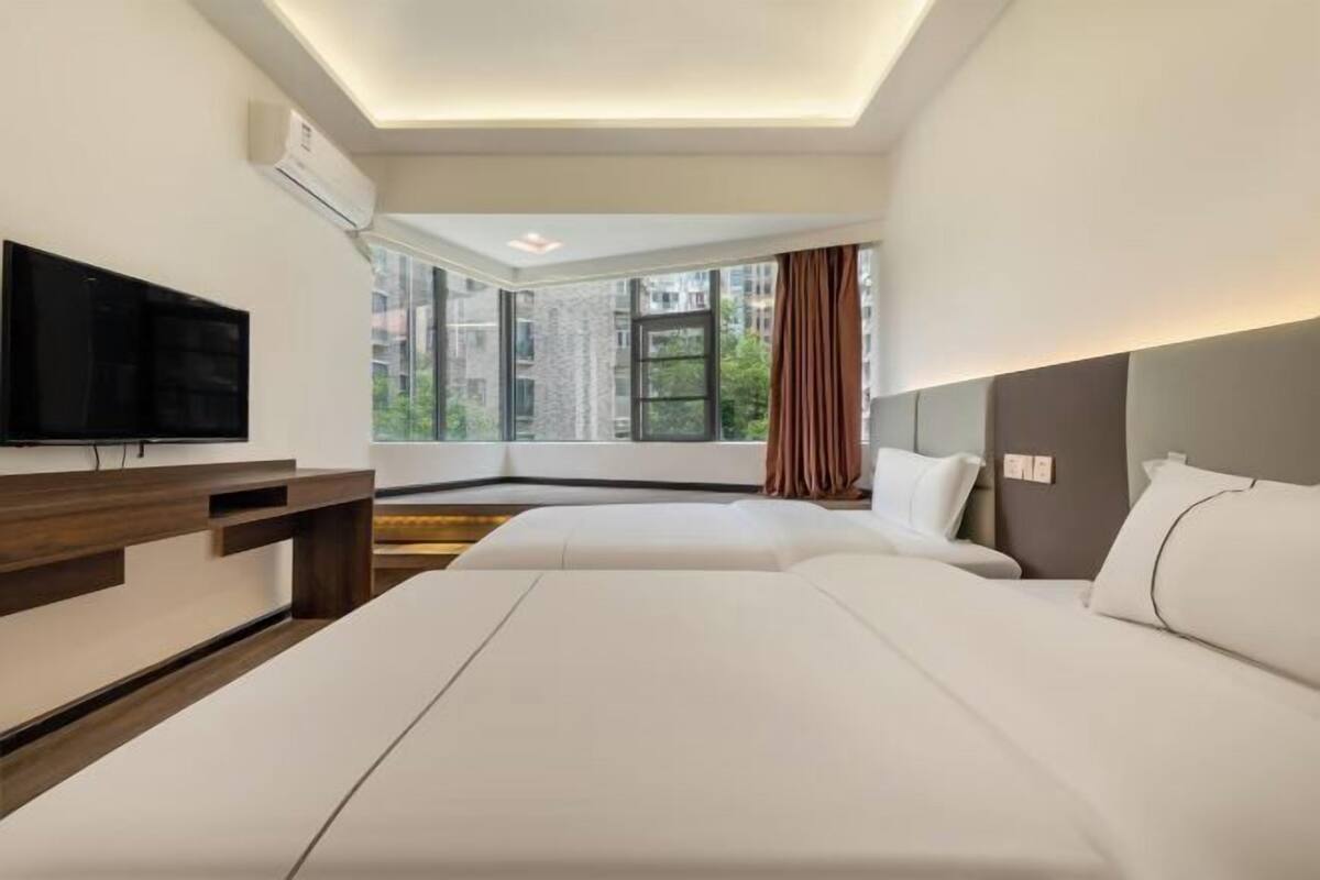 Ein Hotelzimmer mit zwei Betten, einem Flachbildfernseher, einem Holztisch und einem großen Fenster mit Blick auf die Stadt.