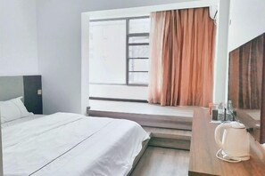 Habitación de hotel con cama, ventana con cortinas, escritorio de madera con hervidor y espejo.