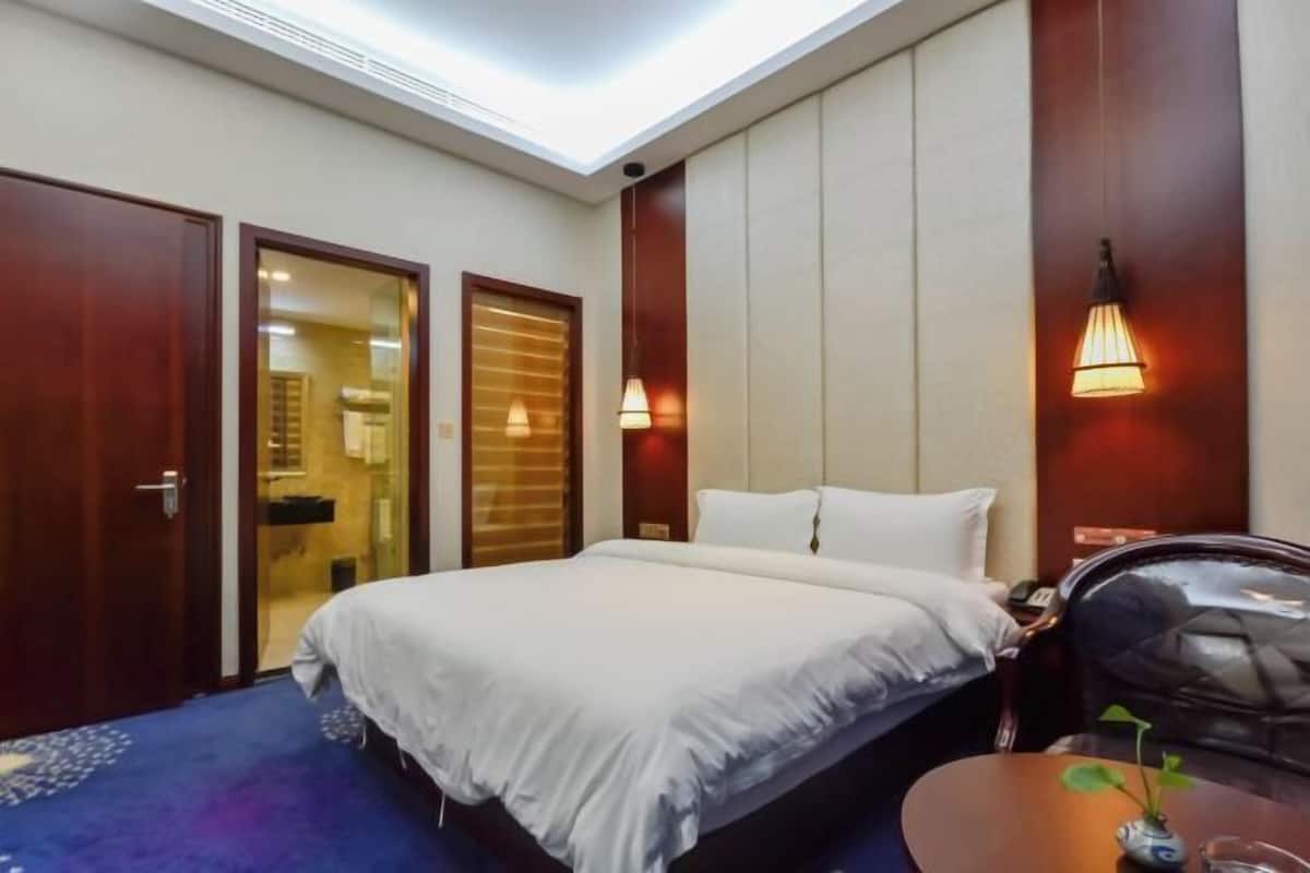 A hotel room with a bed, a desk, a chair, a small table with a plant, and a wall-mounted lamp.