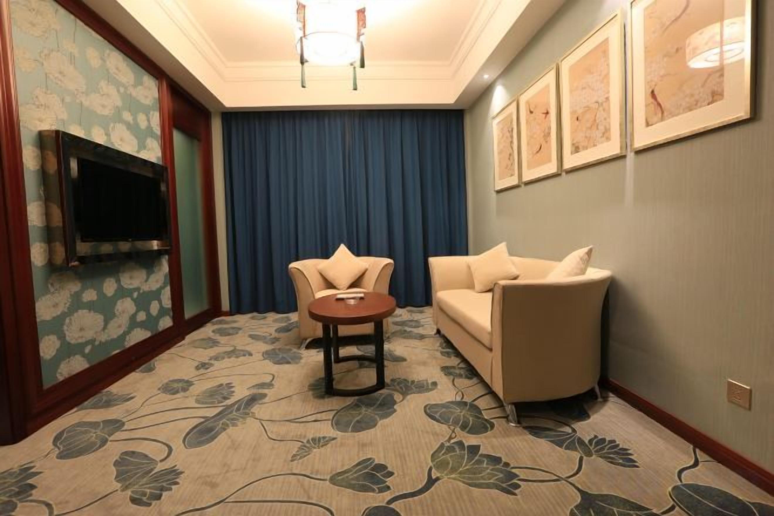 A hotel room with a flat-screen TV, a sofa, a chair, a small table, and a patterned carpet.