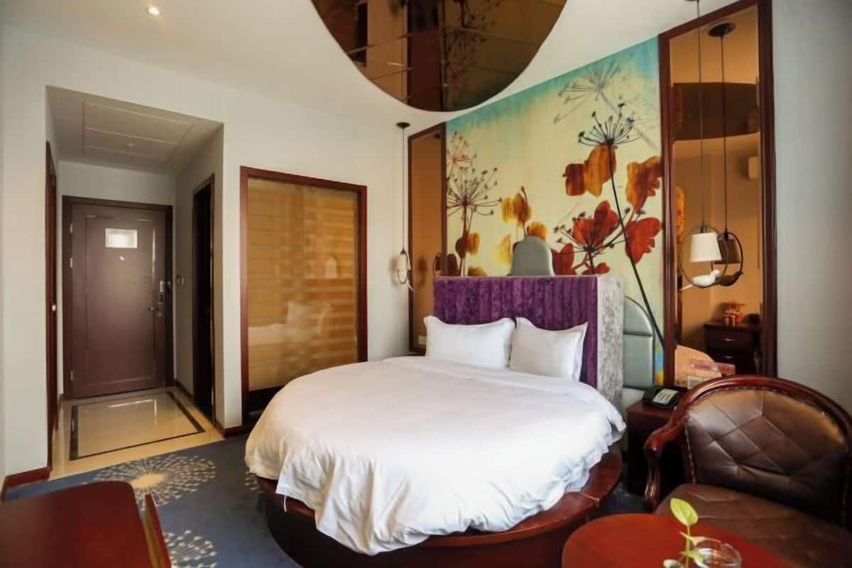 A hotel room with a large bed, a desk, a chair, a door, and a wall with a floral painting.