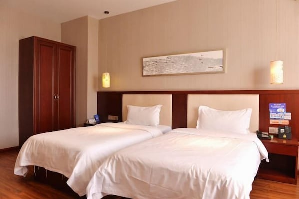 A hotel room with two beds, a wooden wardrobe, a painting on the wall, and a bedside table with a lamp.