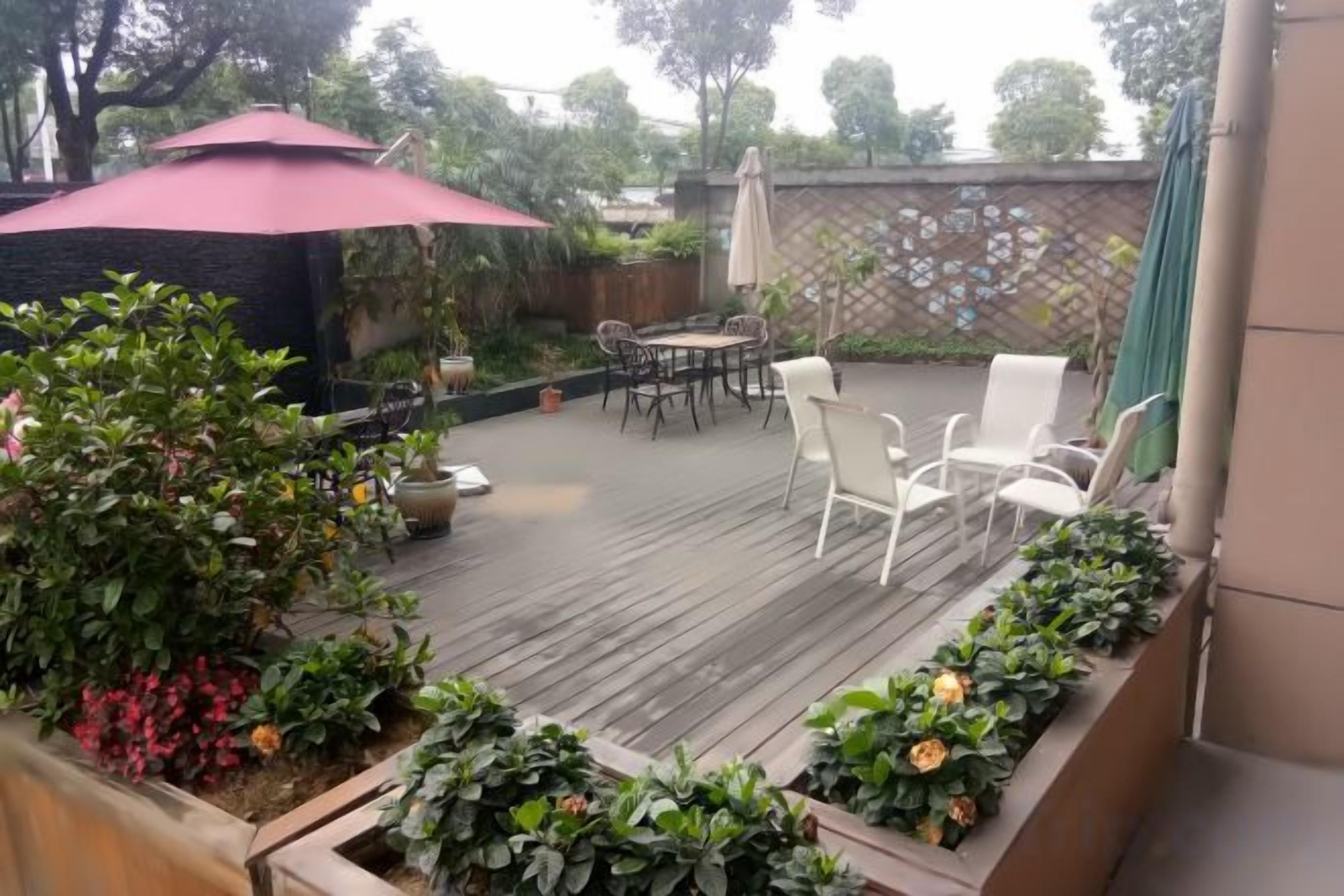 Een houten terras met buitenmeubilair, omringd door potplanten en een rode parasol.