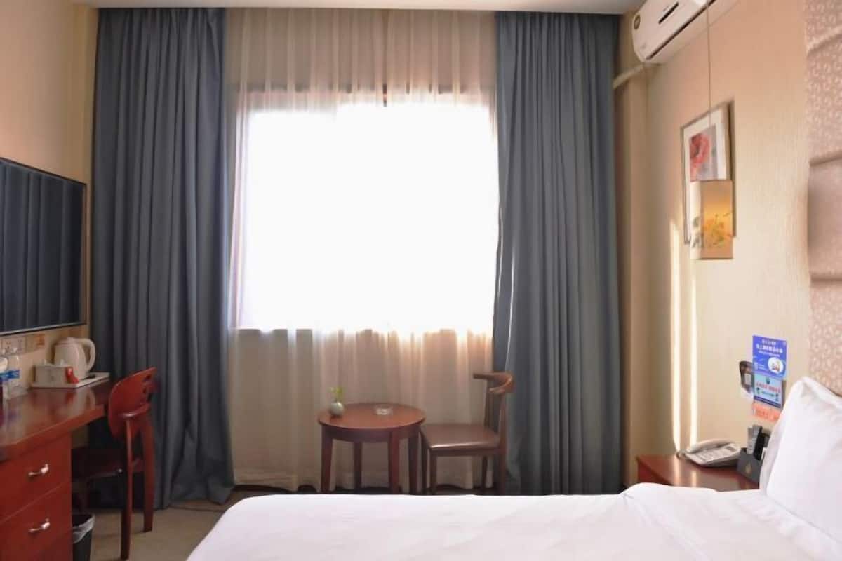 A hotel room with a bed, a desk with a chair, a small table with a chair, a TV, and a window with curtains.