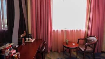 A hotel room with a wooden dining table, two chairs, a small round table with a plant, and a window with pink curtains.