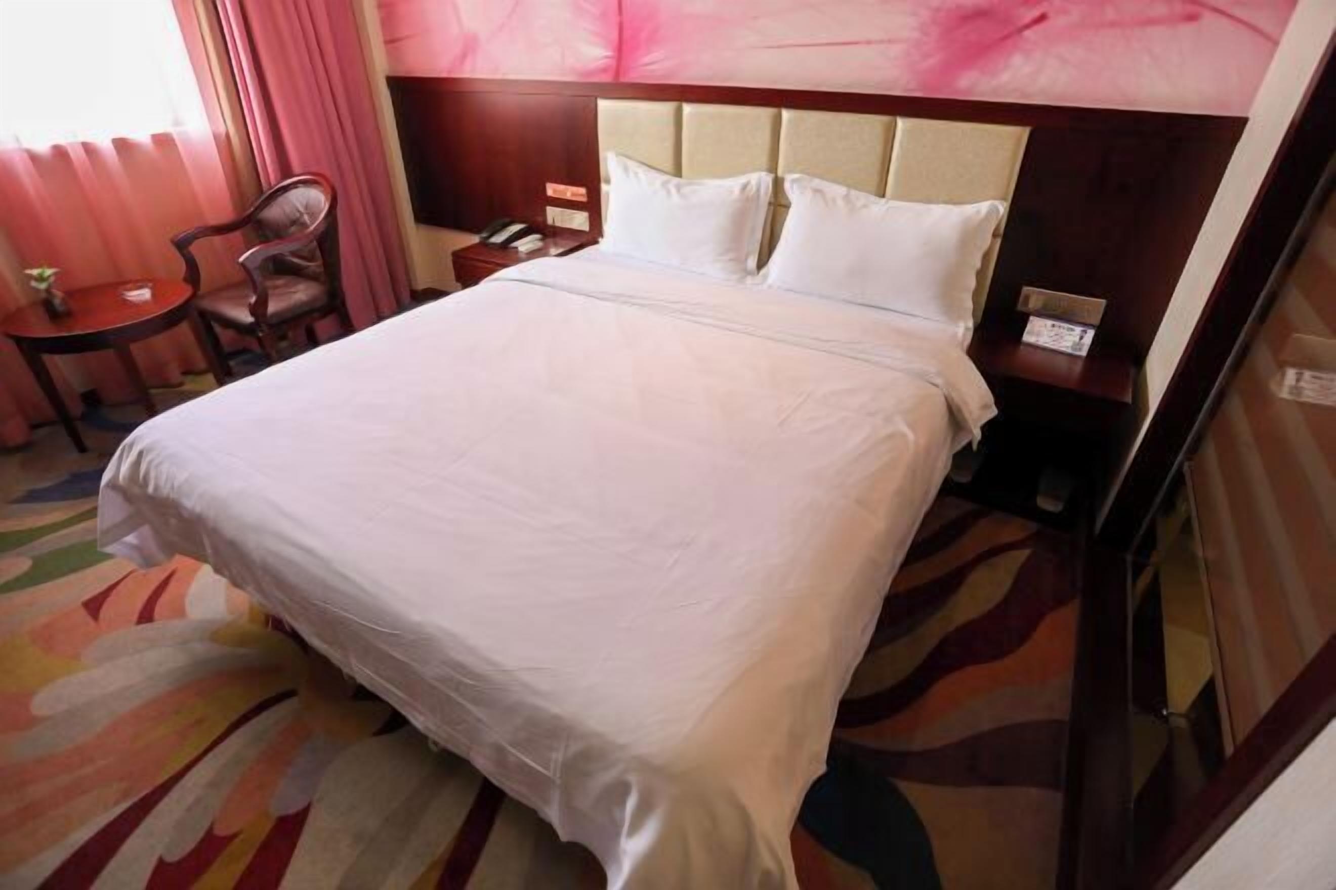 Habitación de hotel con una cama grande, cabecera de madera, mesita de noche con lámpara, una silla y una pequeña mesa con una planta.