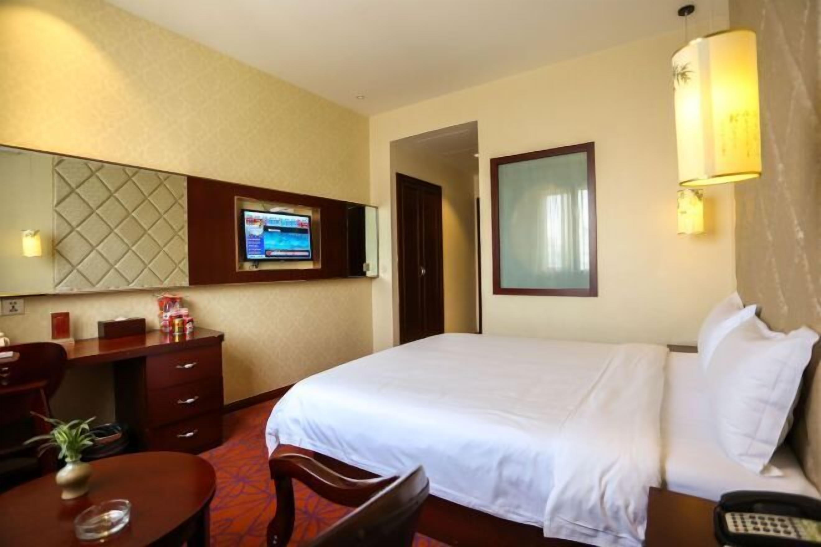 A hotel room with a bed, a desk with a computer, a chair, a small table with a plant, and a telephone.