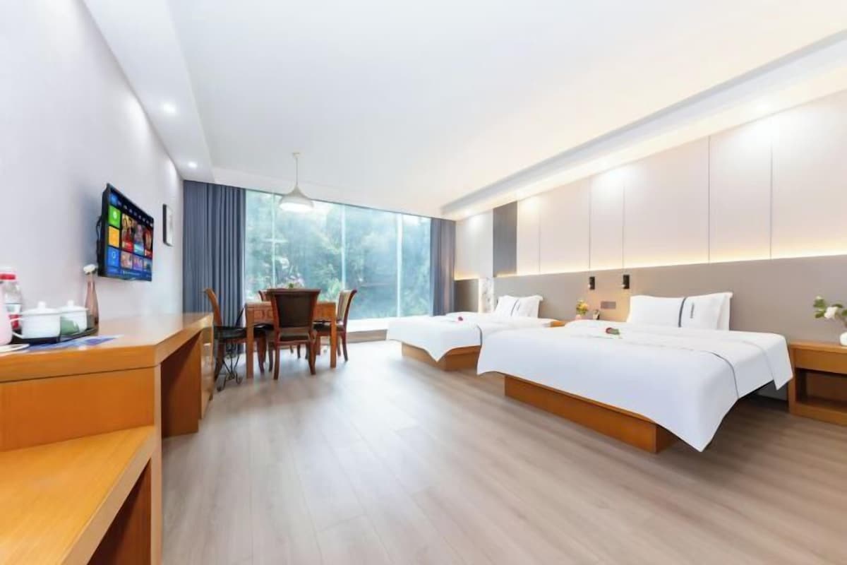 Una habitación de hotel moderna con un escritorio de madera, dos camas, un televisor y un ventanal amplio.