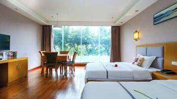 A hotel room with two beds, a dining area with a table and chairs, a TV, and a large window with a view of greenery.