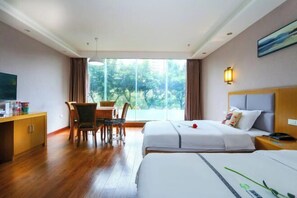 Habitación de hotel con dos camas, un comedor con mesa y sillas, un televisor y un ventanal con vistas a zonas verdes.