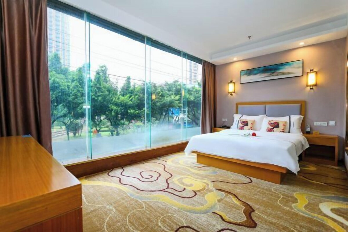 Una habitación de hotel con un ventanal grande, una cama con ropa de cama blanca, un cabecero de madera y una alfombra estampada.